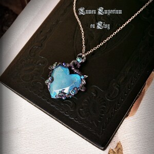 May include: A delicate gold chain necklace with a blue heart-shaped pendant. The pendant is surrounded by intricate black wirework and iridescent green and blue beads.