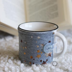 May include: A white ceramic mug with a black rim and handle, wrapped in a gray fabric cozy. The cozy is decorated with gold star patterns and secured with a large, clear button. The mug sits on a white, fluffy surface.