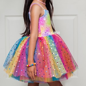 Könnte beinhalten: Ein farbenfrohes Regenbogen-Tutu-Kleid mit einem paillettenbesetzten Oberteil und einem mehrschichtigen Rock. Der Rock ist mit herzförmigen Pailletten verziert. Ein passendes Regenbogen-Armband ist sichtbar. Das Kleid ist für Kinder.