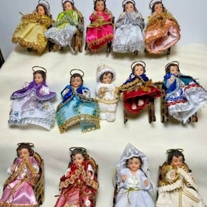 May include: A collection of 17 small, dressed figurines of baby Jesus in various colors and poses. Each figurine is seated on a small wooden chair.