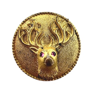 May include: A gold-toned round pin with a detailed deer head design. The deer has large, pink eyes and a textured, sculpted face.