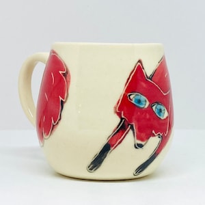 May include: A white ceramic mug featuring a red fox design. The fox has black legs and blue eyes. The mug has a rounded shape and a handle.