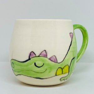May include: A white ceramic mug with a green handle and a hand-painted design of a green alligator with purple spines. The alligator is smiling and has yellow accents. The mug is a rounded shape.