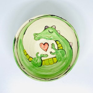May include: A green ceramic bowl with a hand-painted design of a green alligator holding a red heart. The bowl has a white interior and a green rim.