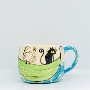 May include: A white ceramic mug with a blue rim and handle. The mug features a green, blue, and white design with a black cat and a white owl.