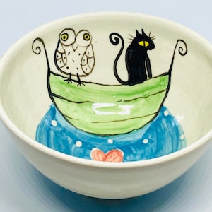 May include: A white ceramic bowl with a hand-painted design of an owl and a black cat in a green boat on a blue and white water background.