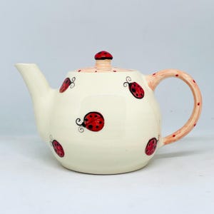 May include: Cream ceramic teapot decorated with red ladybug motifs. The teapot features a curved spout, a rounded body, and a handle adorned with red polka dots. The lid is topped with a red knob. The ladybugs are red with black details.