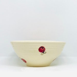 May include: Cream-coloured ceramic bowl with a wide rim and a rounded base. The bowl is decorated with red ladybug illustrations with black spots and curly antennae. The bowl has a rustic, handmade appearance.