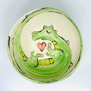 May include: A circular ceramic bowl with a hand-painted design. The bowl features a green cartoon alligator encircling the interior, with a red heart in the centre. The alligator has purple spikes and yellow accents. The bowl's rim is cream-coloured.