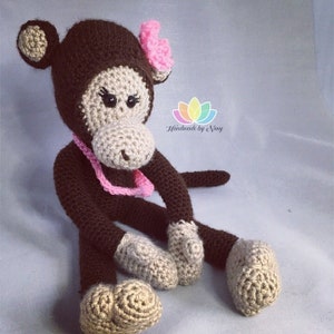 May include: A handmade crochet monkey doll in brown, beige, and pink. The monkey features a brown head and body, beige face, hands, and feet, and a pink flower and necklace. The doll is sitting with its legs extended. The text "Handmade by Niny" is visible.