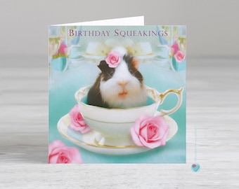 May include: A birthday card featuring a black and white guinea pig wearing a pink flower in its hair, sitting in a teacup with a pink rose on the side. The card has the text "Birthday Squeakings" on the front.