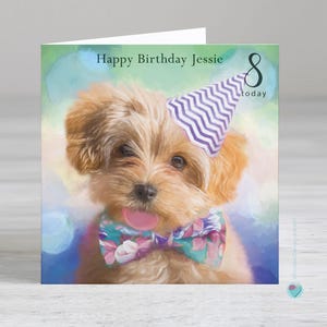 May include: A birthday card featuring a cute, fluffy, light brown puppy wearing a purple and white striped party hat and a colorful bow tie. The card says "Happy Birthday Jessie 8 today".