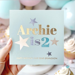 May include: A pale blue birthday card with the words "Archie is 2" in gold and teal lettering, decorated with star accents. The card reads "Happy Birthday Our Dear Grandson" in gold. Cupcakes and balloons are in the background.