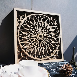 Peut inclure: Une décoration murale en bois avec un motif géométrique en forme de cercle. Le motif est composé de plusieurs couches de bois, créant un effet 3D. La décoration est de couleur brun clair et possède un cadre carré.