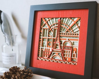 Könnte beinhalten: Ein gerahmtes Holzbild mit rotem Hintergrund und einem geschichteten Design des Eiffelturms und Gebäuden in Paris. Das Kunstwerk befindet sich in einem schwarzen Rahmen.