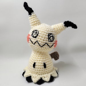 May include: A crocheted plush toy of a Ghost-type Pokemon, Gengar, with a white body, black eyes, and a brown tail. The toy has a friendly expression and is standing on a white background.