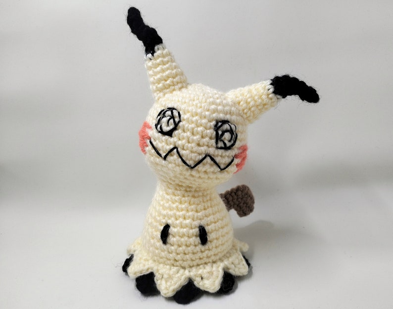 May include: A crocheted plush toy of a Ghost-type Pokemon, Gengar, with a white body, black eyes, and a brown tail. The toy has a friendly expression and is standing on a white background.