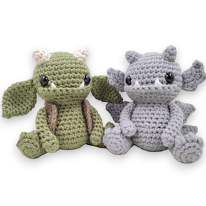 May include: Two crocheted plush toys shaped like small dragons. One is green with white horns and the other is gray with white horns.