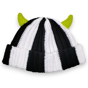 May include: A handmade crochet beanie with alternating black and white vertical stripes. The hat features two bright green horns on the top. The brim is folded up. This unique hat is perfect for costume parties or everyday wear.