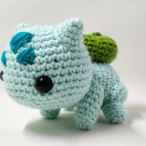 May include: A crocheted green and blue Bulbasaur plush toy. The toy has a green leaf on its back and black eyes.