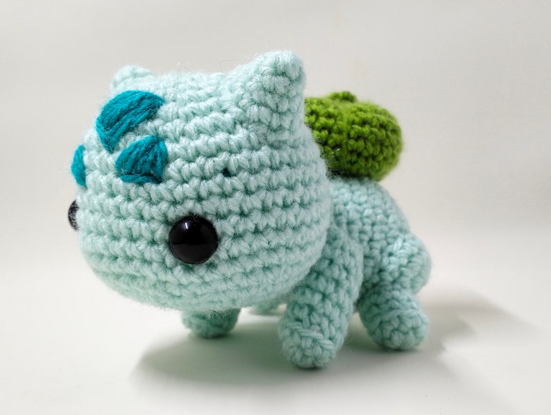 May include: A crocheted green and blue Bulbasaur plush toy. The toy has a green leaf on its back and black eyes.