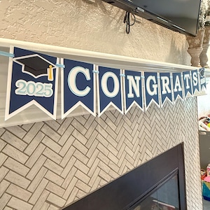 May include: A blue and white graduation banner with the word "CONGRATS" and the year "2025" printed on it. The banner is hanging above a fireplace.