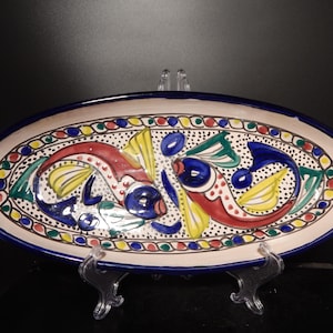 Könnte beinhalten: Eine ovale Keramikplatte mit einem handbemalten Design aus zwei stilisierten Fischen in Rot, Blau, Gelb und Grün. Der Hintergrund ist cremefarben mit schwarzen Sprenkeln und der Rand ist mit einem bunten Kettengliedmuster in Blau verziert.