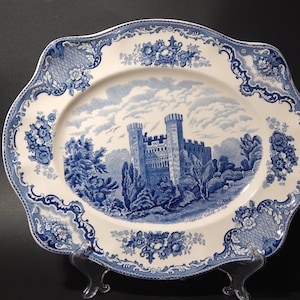 Puede incluir: Plato ovalado de cerámica con un diseño azul y blanco. El centro presenta una escena de castillo, rodeada de bordes florales y de volutas. El plato está sostenido por un soporte transparente.