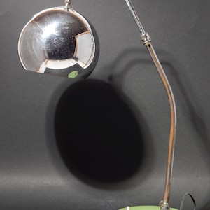 Op de afbeelding: Een chromen bureaulamp met een grote, ronde, chromen kap en een groene voet. De lamp heeft een flexibele arm en een enkele gloeilamp.