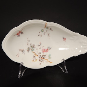 Könnte beinhalten: Eine weiße Porzellanservierschale mit einem Blumendesign in Rosa-, Blau- und Grüntönen. Die Schale ist oval geformt mit einem gewellten Rand und einem erhöhten Rand.