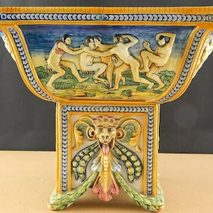 May include: A ceramic bowl with a yellow, blue and green design. The bowl features a scene of five nude figures dancing. The bowl sits on a pedestal with a grotesque face and floral designs.