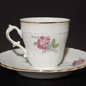 Puede incluir: Un juego de taza y platillo de té de porcelana blanca con ribete dorado y diseño floral. La taza tiene un asa delicada y el platillo tiene un borde festoneado.