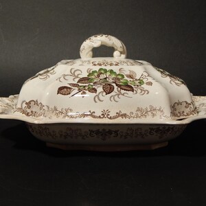 Può includere: Un piatto da portata in ceramica bianca con coperchio. Il piatto ha un motivo floreale marrone e verde. Il coperchio ha un manico.