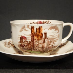 Può includere: Una tazza da tè e un piattino bianchi con un disegno di castello marrone e rosso. Il castello è circondato da alberi e acqua.