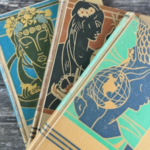 Pode incluir: Uma pilha de livros vintage com ilustrações em estilo art déco. As capas apresentam imagens de mulheres e um globo, em tons de azul, verde, castanho e dourado. Os livros têm lombadas douradas.