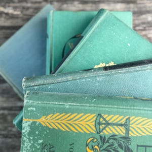 Puede incluir: Una pila de libros antiguos con cubiertas en tonos turquesa y azul. Los libros tienen bordes desgastados y letras doradas. Uno de los libros presenta un dise&ntilde;o de pluma amarilla. Los libros est&aacute;n dispuestos en &aacute;ngulo, mostrando sus diferentes tonos y texturas.