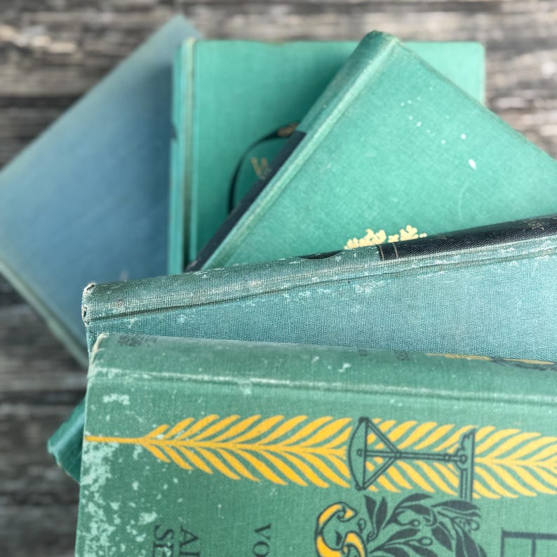 Puede incluir: Una pila de libros antiguos con cubiertas en tonos turquesa y azul. Los libros tienen bordes desgastados y letras doradas. Uno de los libros presenta un dise&ntilde;o de pluma amarilla. Los libros est&aacute;n dispuestos en &aacute;ngulo, mostrando sus diferentes tonos y texturas.
