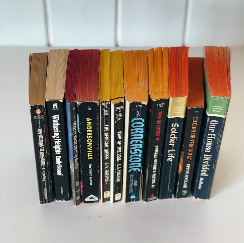 May include: A collection of 12 paperback books with various titles and authors. The books are arranged vertically in a row, with the spines facing the viewer. The books are all different colors, including red, orange, yellow, blue, and green.