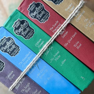 Op de afbeelding: Een stapel klassieke boeken, gebonden in verschillende kleuren, vastgebonden met touw. De ruggen tonen titels als "De Scharlaken Pimpernel", "Tom Sawyer" en "Schatteneiland". Elk boek heeft het logo "Reader's Digest Best Loved Books".