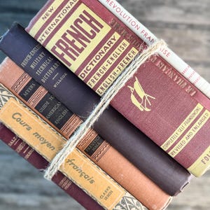 May include: A stack of five vintage hardcover books tied together with rustic twine. The collection includes a New International French Dictionary, a French-English military technical dictionary, and various language textbooks with titles like Cours moyen.