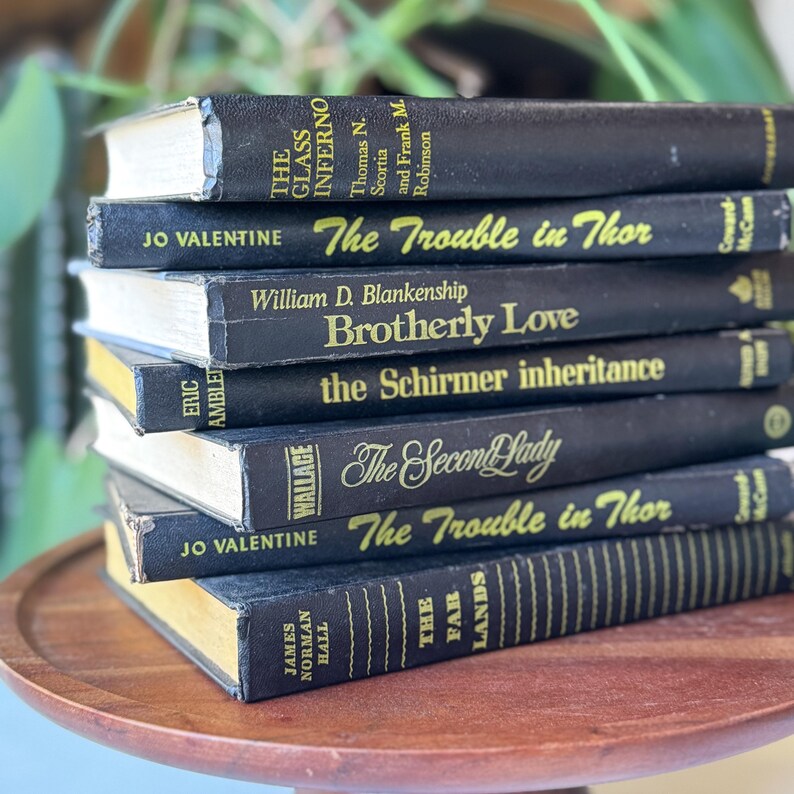 May include: A stack of vintage hardcover books with black covers and gold lettering. Titles include "The Glass Inferno," "The Trouble in Thor," and "Brotherly Love." The books are arranged on a wooden tray.