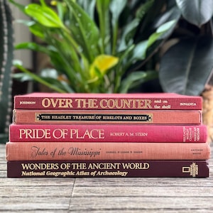 Könnte beinhalten: Ein Stapel von fünf alten Büchern mit roten und kastanienbraunen Einbänden. Die Titel sind unter anderem "Over the Counter" und "Pride of Place". Die Bücher sind auf einer Holzoberfläche gestapelt, mit grünen Pflanzen im Hintergrund.