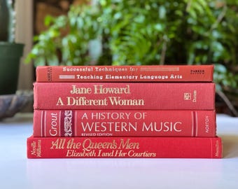 May include: A stack of four vintage books with red covers. Titles include "Successful Techniques for Teaching Elementary Language Arts," "A Different Woman," "A History of Western Music," and "All the Queen's Men." The books are stacked on a white surface with green foliage in the background.