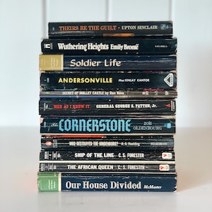May include: A stack of 11 black paperback books with white text titles. The titles include "Their Be The Guilt", "Wuthering Heights", "Soldier Life", "Andersonville", "Secret of Mallet Castle", "War As I Knew It", "The Cornerstone", "Who Destroyed The Hindenburg", "Ship of the Line", "The African Queen", and "Our House Divided".