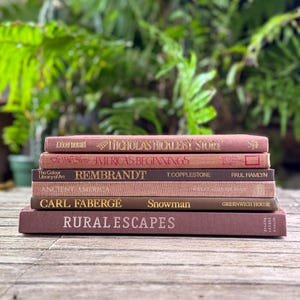 May include: A stack of six hardcover books with fabric covers in shades of dusty rose, mauve, and brown. The spines feature gold and white text including titles like The Nicholas Nickleby Story, America's Beginnings, Rembrandt, Ancient America, Carl Fabergé Snowman, and Rural Escapes.