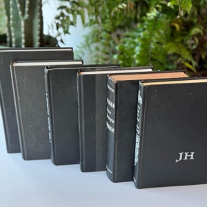 May include: A row of six black hardcover books with white lettering on the spines. The books are arranged in order of size, with the largest book on the left and the smallest book on the right. The books are all closed and have a smooth, glossy finish.
