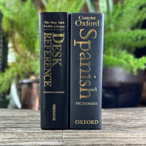 Peut inclure: Deux livres à couverture rigide noirs sont présentés. Le livre de gauche est intitulé "DESK REFERENCE" de la New York Public Library. Le livre de droite est un "Concise Oxford Spanish Dictionary". Les deux livres ont des lettres dorées.