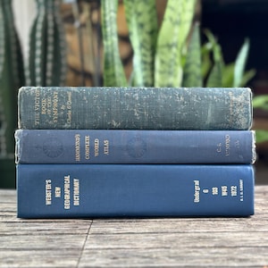 Puede incluir: Tres libros antiguos apilados sobre una superficie de madera. El libro superior es verde azulado, titulado "The Victor Book of the Symphony". El libro del medio es azul, titulado "Hammond's Complete World Atlas". El libro inferior es azul, titulado "Webster's New Geographical Dictionary".