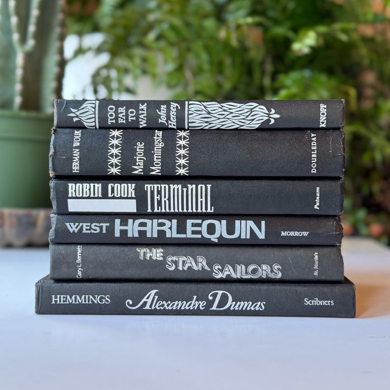 May include: A stack of black hardcover books with white text titles. The titles include "Too Far to Walk", "Terminal", "Harlequin", "The Star Sailors", and "Alexandre Dumas".