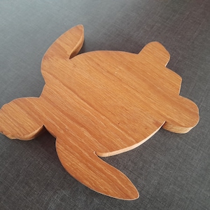 Puede incluir: Una tabla de cortar de madera con forma de tortuga, con un acabado de vetas de madera natural. La tabla es de color marrón claro y tiene una superficie lisa. La cabeza, las aletas y la cola de la tortuga están claramente definidas. Este artículo decorativo es adecuado para la cocina.